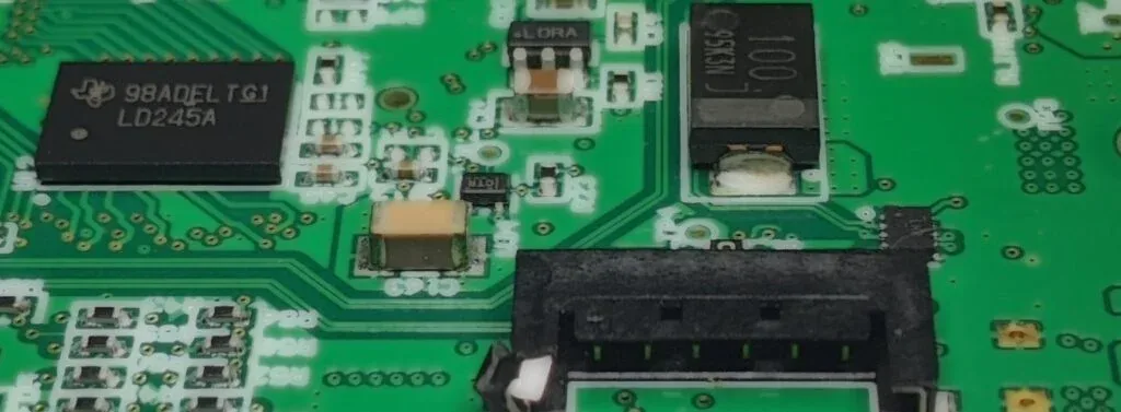 Gros plan d'une carte électronique d'un produit IoT compact conçu par AESTECHNO en 2020 : contrôleur Texas Instruments, module radio LoRa sub-GHz (silkscreen LORA visible), oscillateur quartz NX35 et connecteur batterie JST.